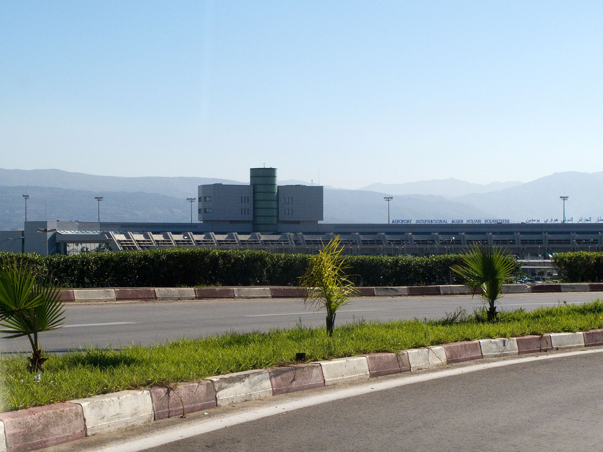 A long low airport terminal with an airport name sign and a control tower block, seen across a road and palm plantings with hills behind under a clear sky