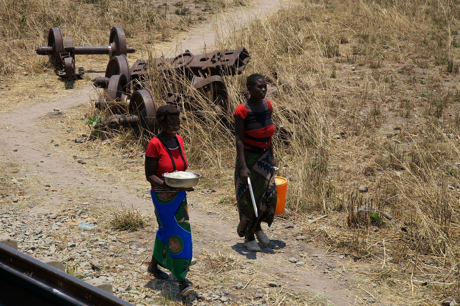 Two women in colourful dresses carry a bowl of food beside a dirt path next to the TAZARA railway line, with old wheelsets in the grass
