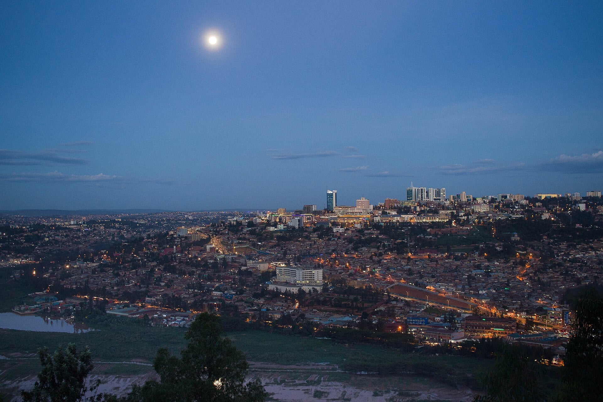 Kigali at dusk seen from a hillside, with city lights across the hills, a cluster of office towers and a full moon in a blue sky