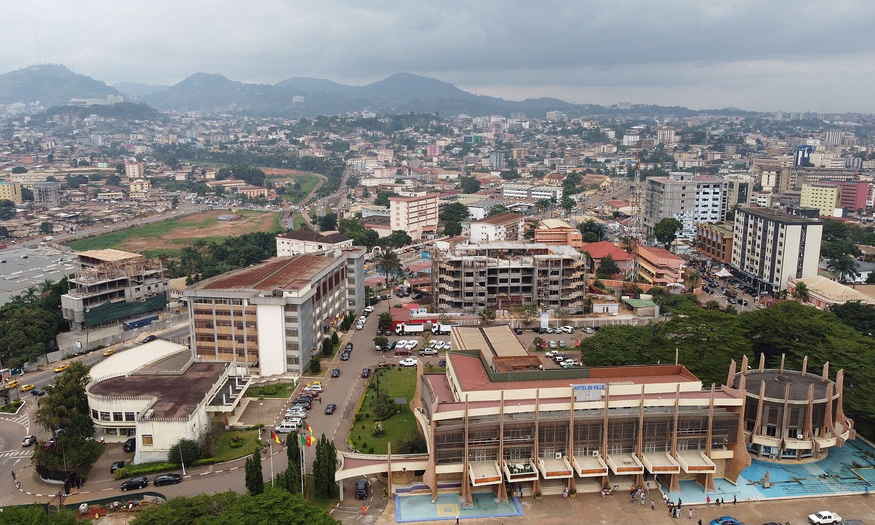 Aerial view of central Yaoundé, Cameroon, with government and commercial buildings, tree-lined streets and green hills on the horizon under an overcast sky