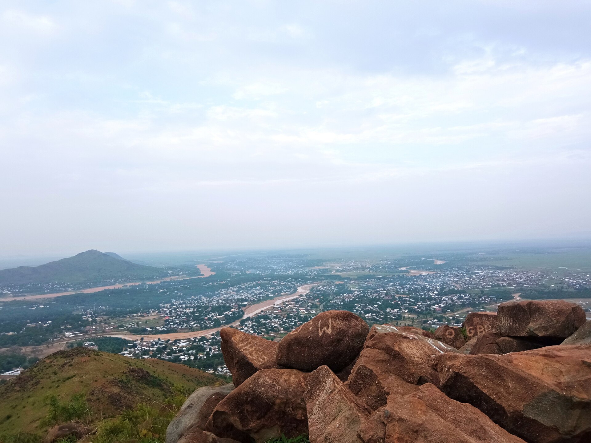 View over the city of Maroua and a winding river bed from red granite boulders on Mont Mayo Ibé, with a hill on the left and a hazy sky