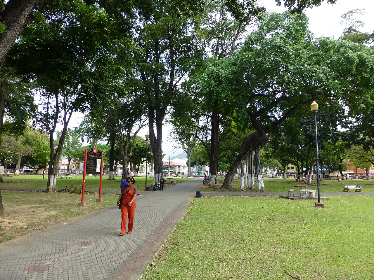 Visitors walk a paved path under large shade trees in Woodford Square, a public park in central Port of Spain.
