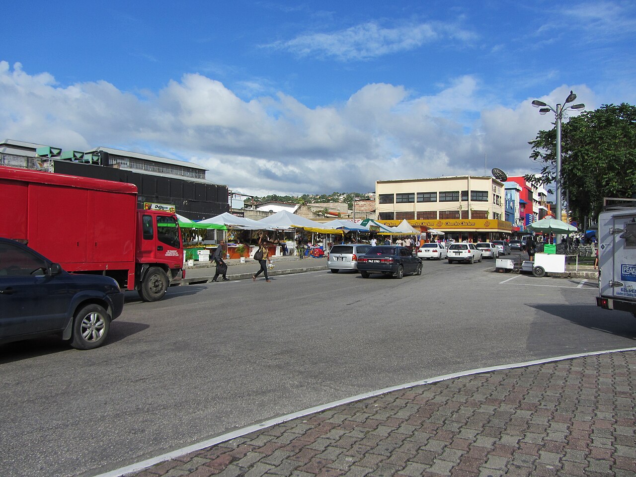 Cars and market stalls on Independence Street in central Port of Spain under a blue sky.