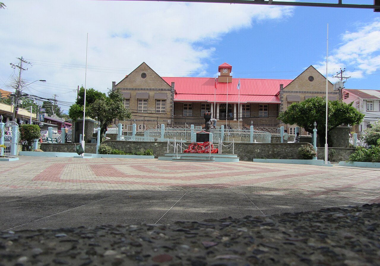 A red-roofed brick building with a clock tower behind a paved public square and war memorial in Scarborough, Tobago.