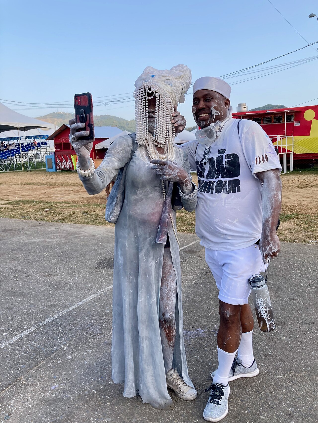 Two revellers covered in white powder and pearls take a phone photo at a jouvay Carnival gathering in Trinidad, with hills behind