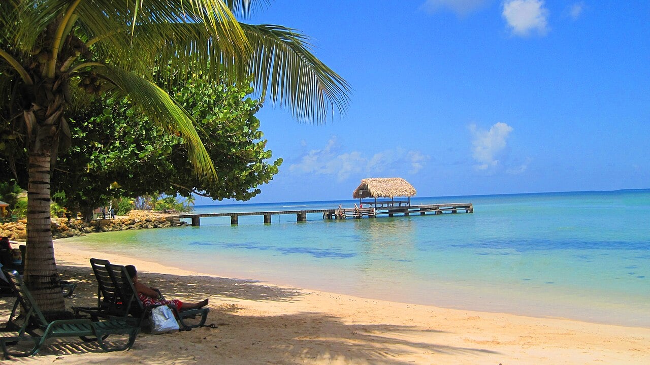 A wooden pier with a thatched hut over turquoise water at Pigeon Point beach in Tobago, with a palm tree and sun loungers on the sand