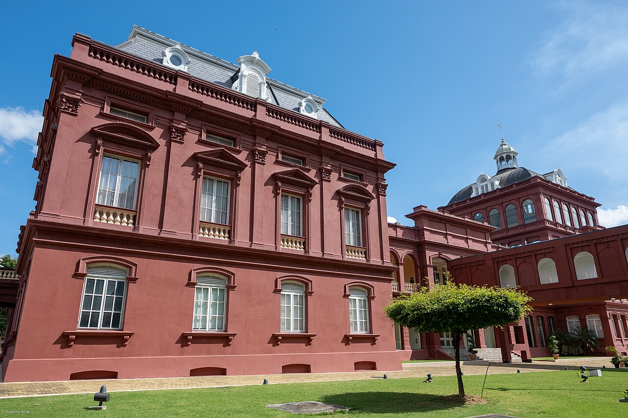 Side view of the red Red House parliament building in Port of Spain with a mansard roof, a small dome in the background and a lawn with a tree