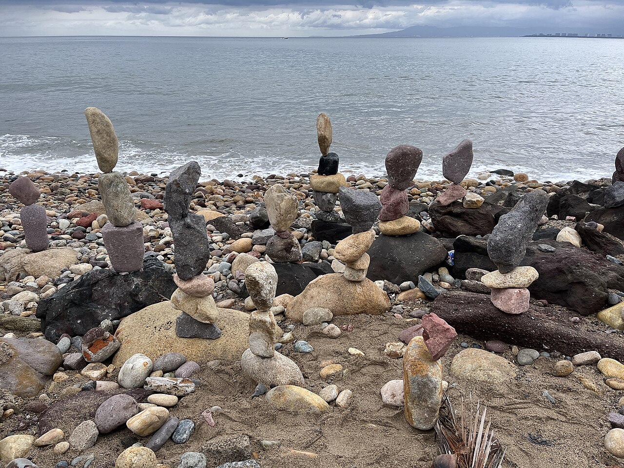 Stacked balancing stones on a pebble beach in Puerto Vallarta under a cloudy sky, with the water of Banderas Bay behind