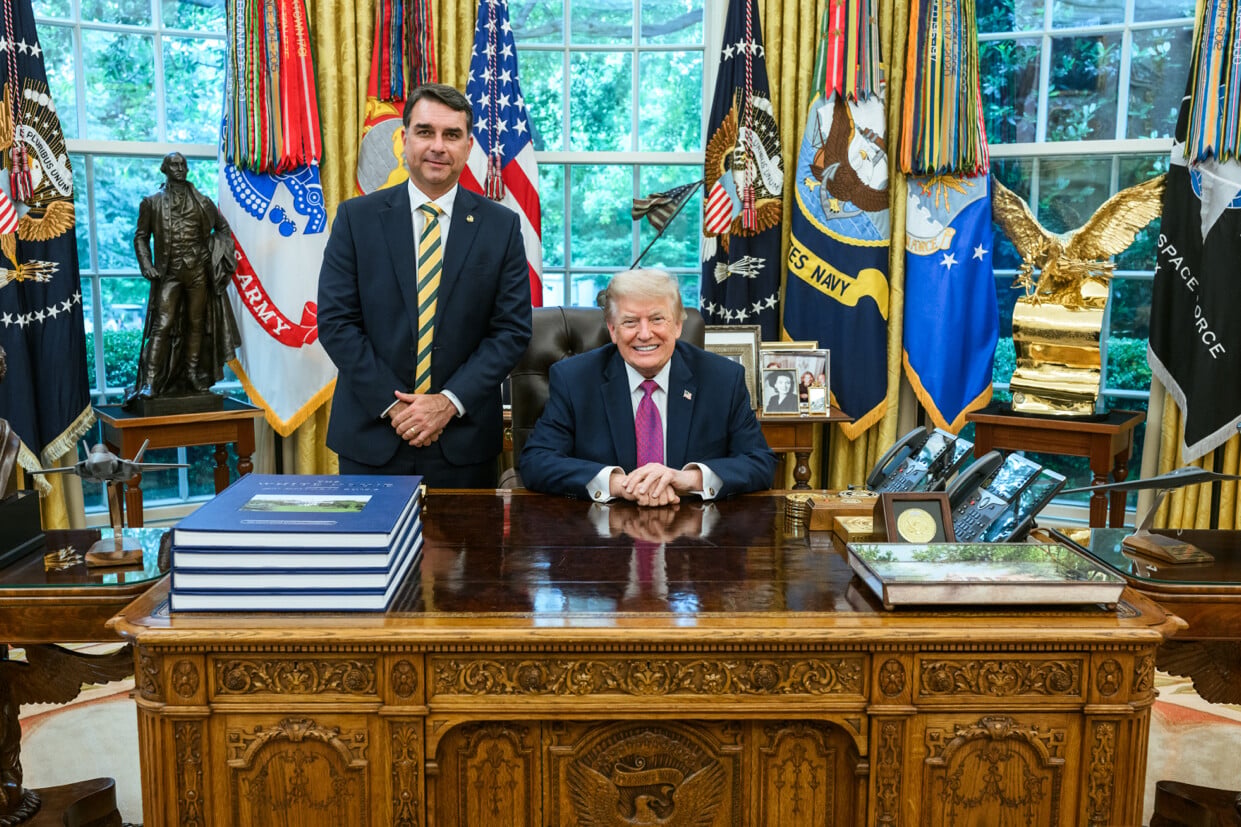 President Donald Trump seated at the Resolute Desk with Brazilian Senator Flávio Bolsonaro standing beside him in the Oval Office