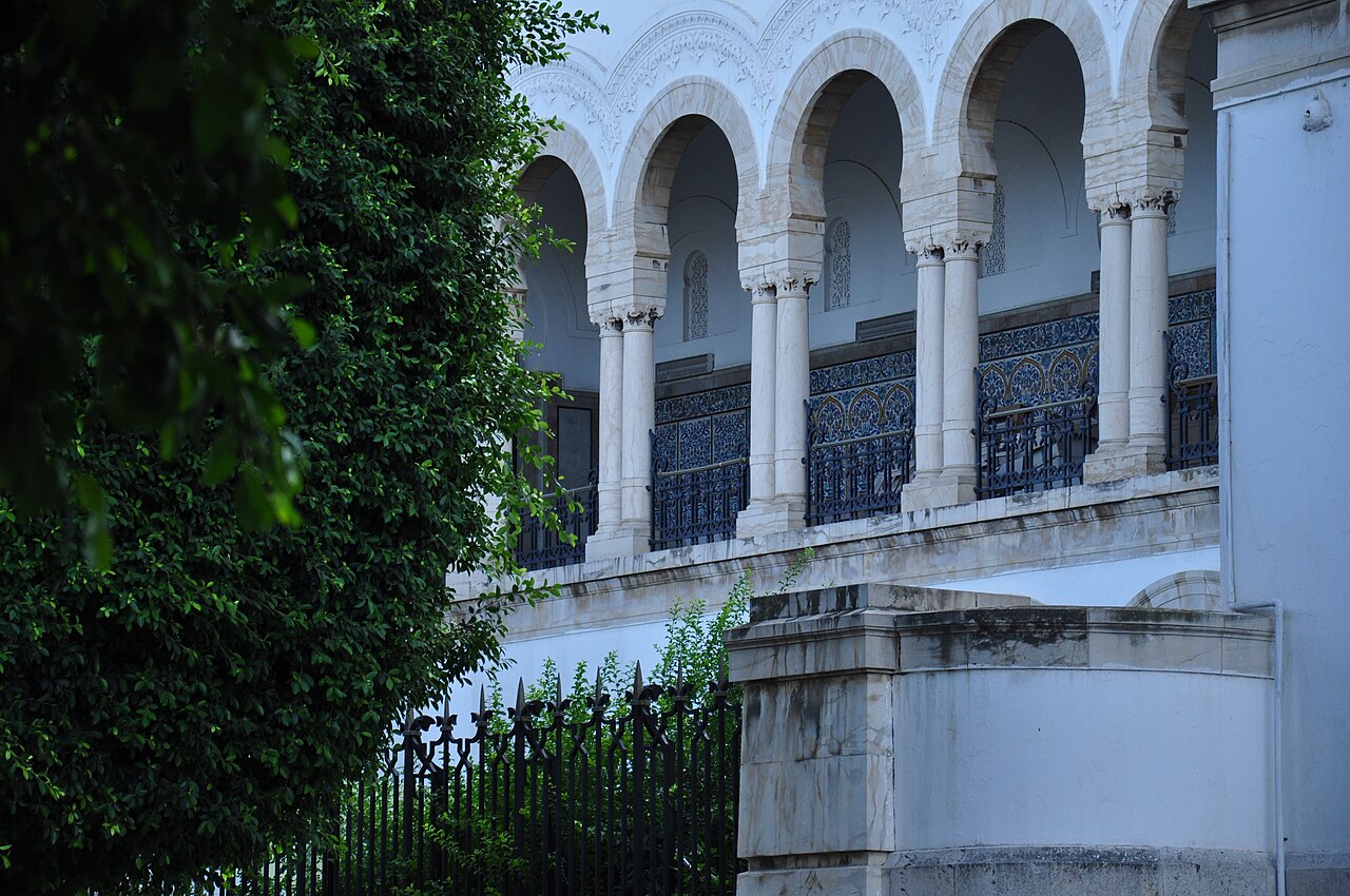 Stone arches, slender columns and dark blue wrought-iron railings on an upper gallery of a white building in Tunis, with green foliage in front