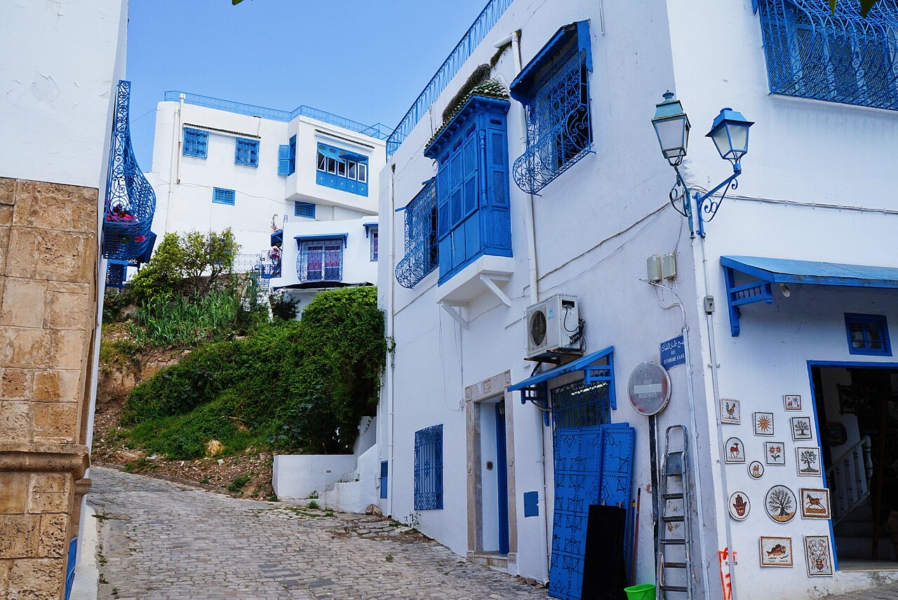 Narrow cobbled lane in Sidi Bou Said, Tunisia, with white houses, blue wooden balconies and blue doors and a shop with small ceramic tiles