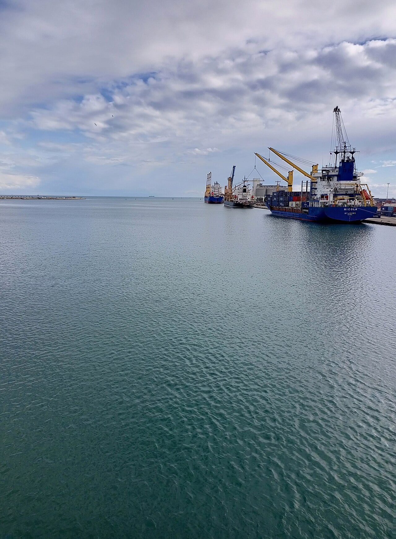 Cargo ships with yellow cranes moored at a quay in the port of Sfax under a cloudy sky, with calm green water in the foreground.