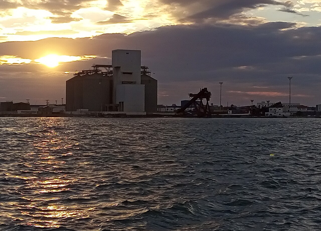 The dark outline of a large grain silo and a loading crane at the port of Sfax at sunset, seen across rippling water.