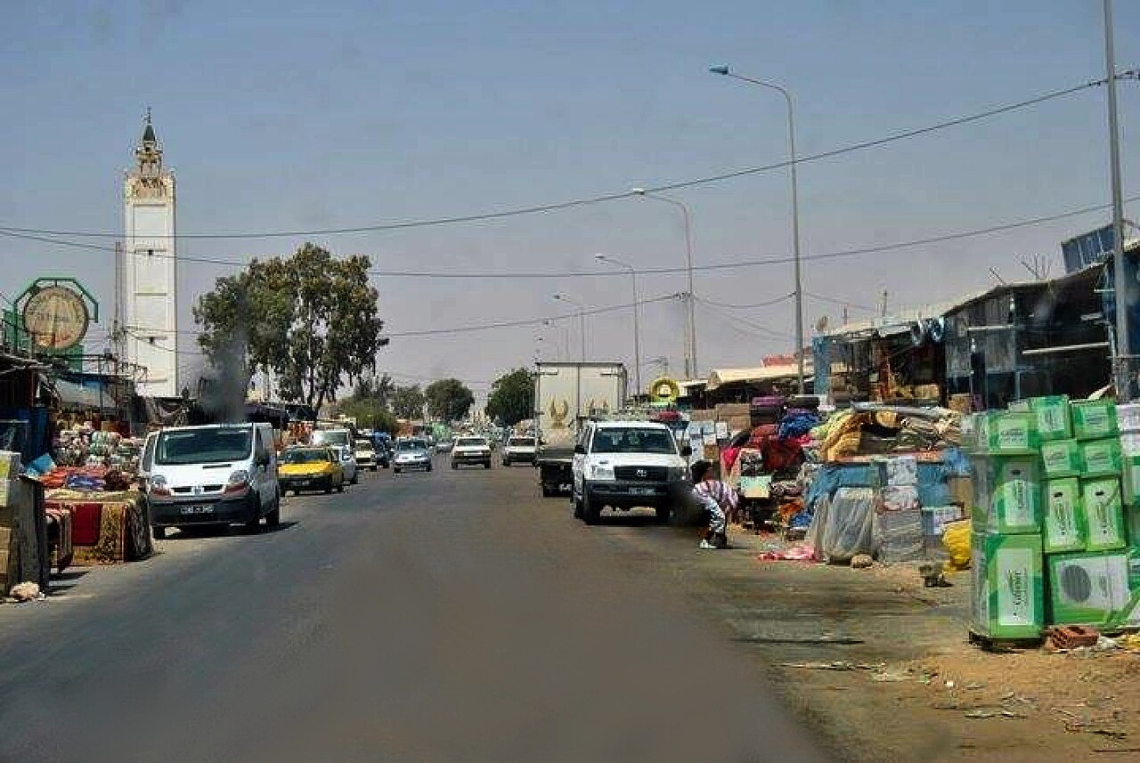 A busy road in the centre of Ben Gardane, with a white minaret, market stalls piled with carpets and boxed goods on both sides, and vans and cars driving down the street.