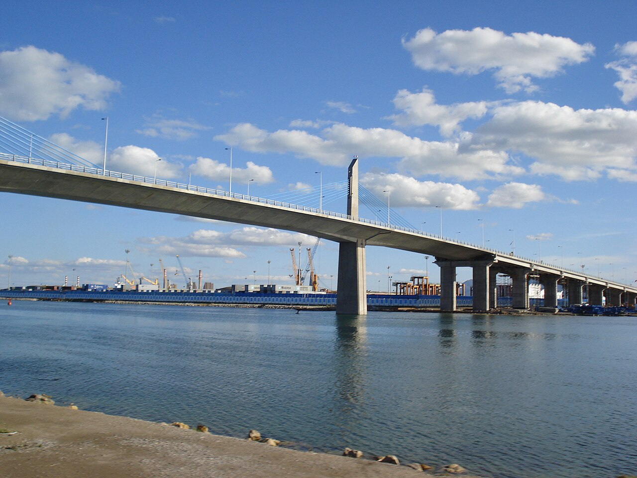 A long concrete cable-stayed bridge over calm water, with container cranes and stacked containers of a port behind it under a blue sky.