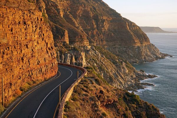 Winding coastal road cut into an orange cliff above the sea on Chapman's Peak Drive near Cape Town, in warm evening light.