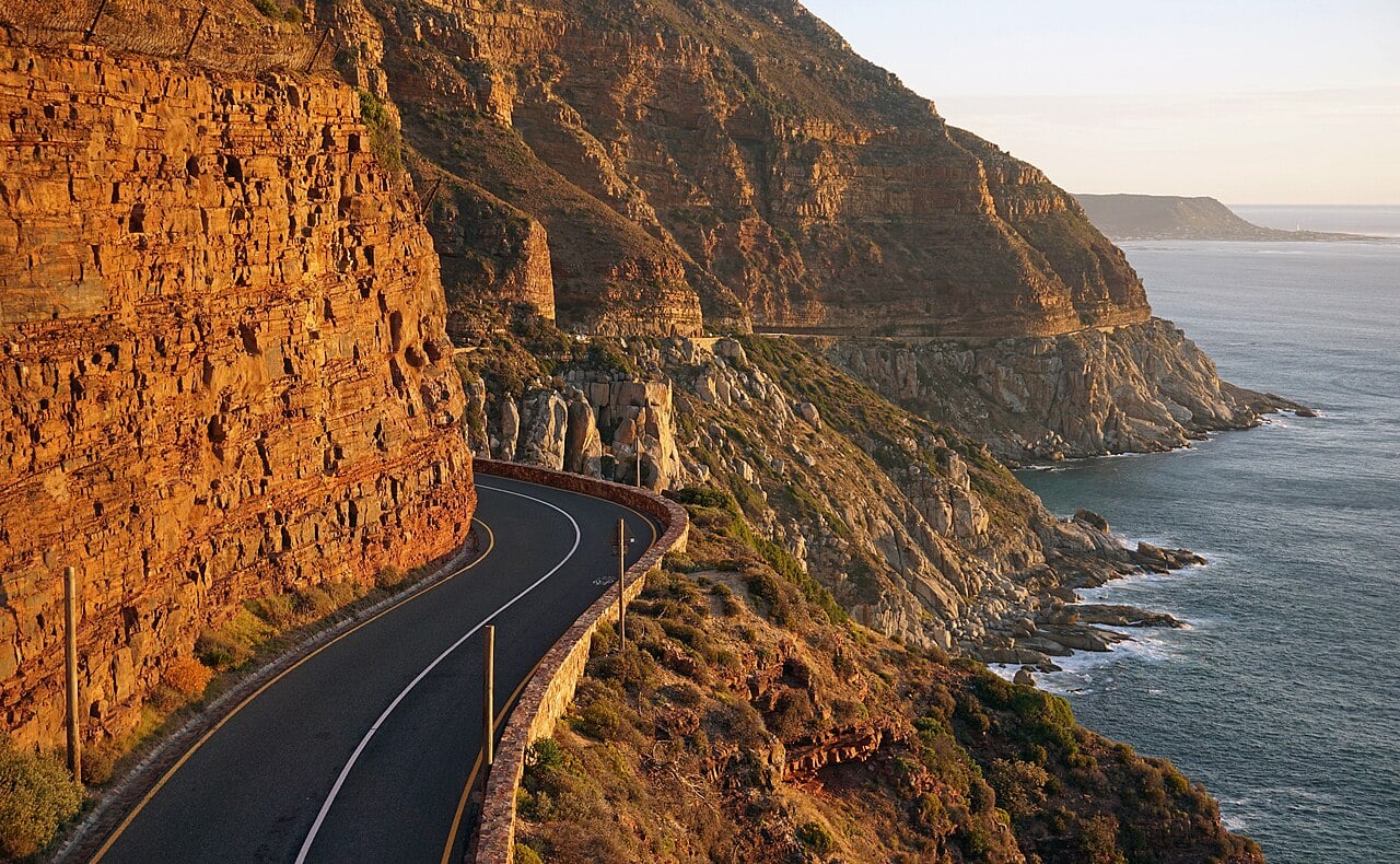 Winding coastal road cut into an orange cliff above the sea on Chapman's Peak Drive near Cape Town, in warm evening light.
