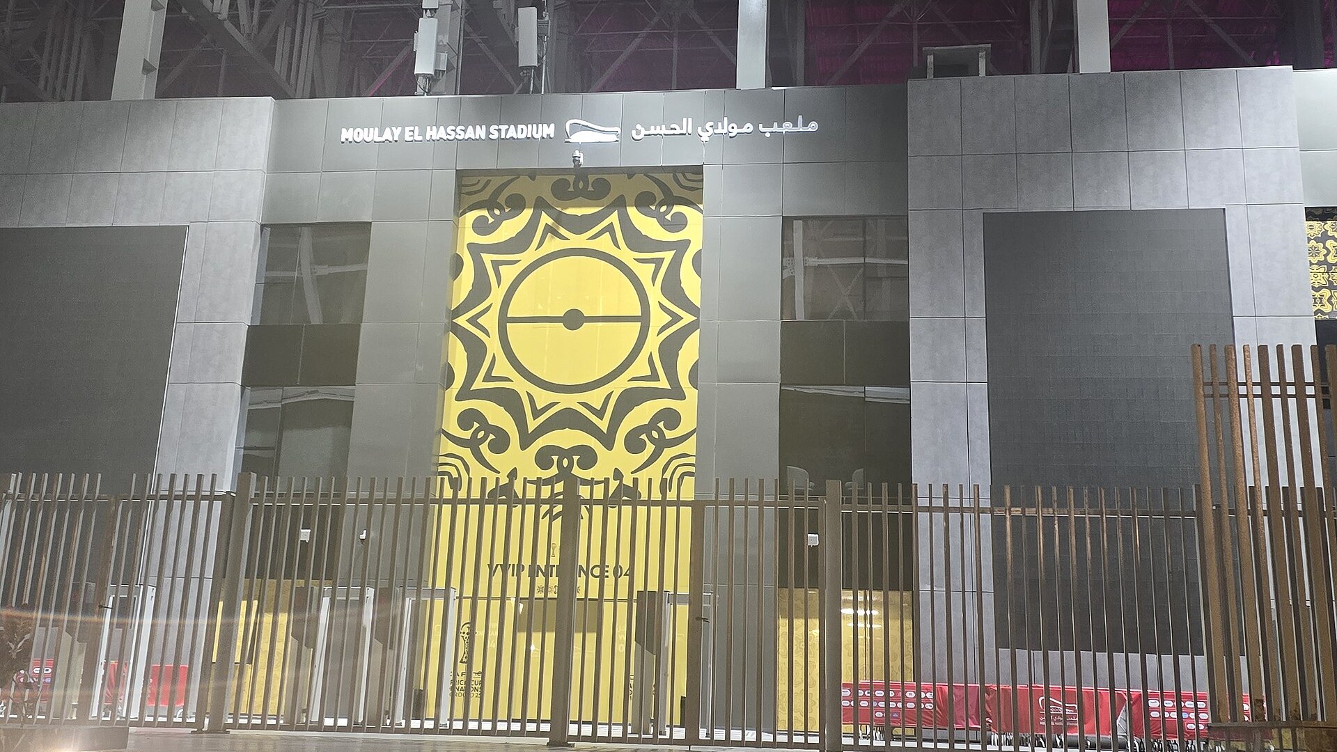 Illuminated entrance of Moulay El Hassan Stadium in Rabat at night, with the stadium name in English and Arabic above a gold patterned gate