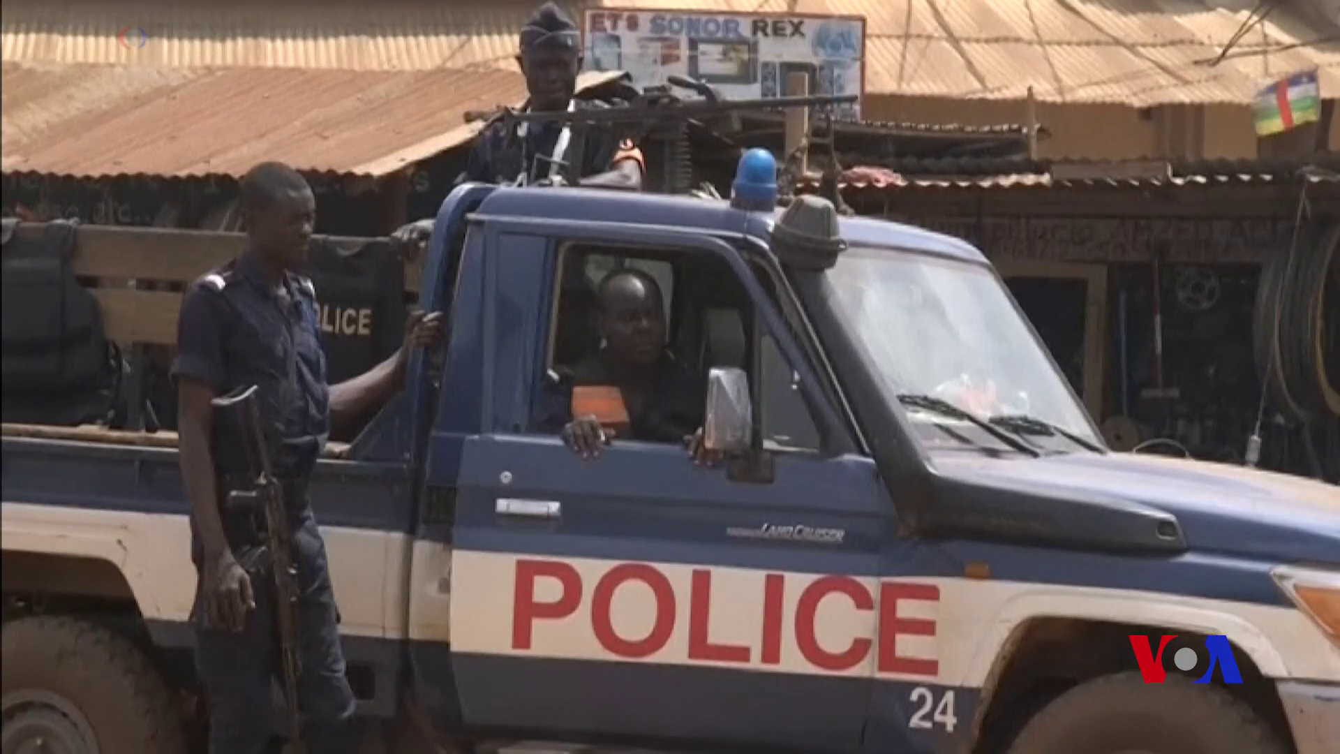 Two Central African police officers, one armed and standing beside a blue and white police pickup, another manning a mounted gun on the back, in a Bangui street