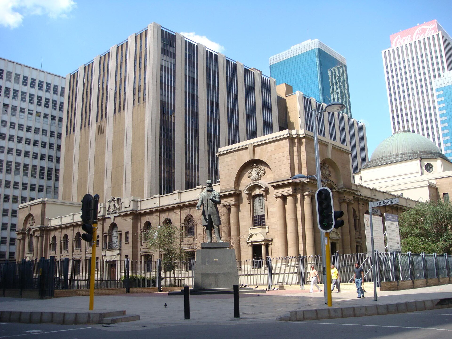 Sandstone court building with arched entrance and a bronze statue in front, office towers behind, on a sunny street in central Johannesburg