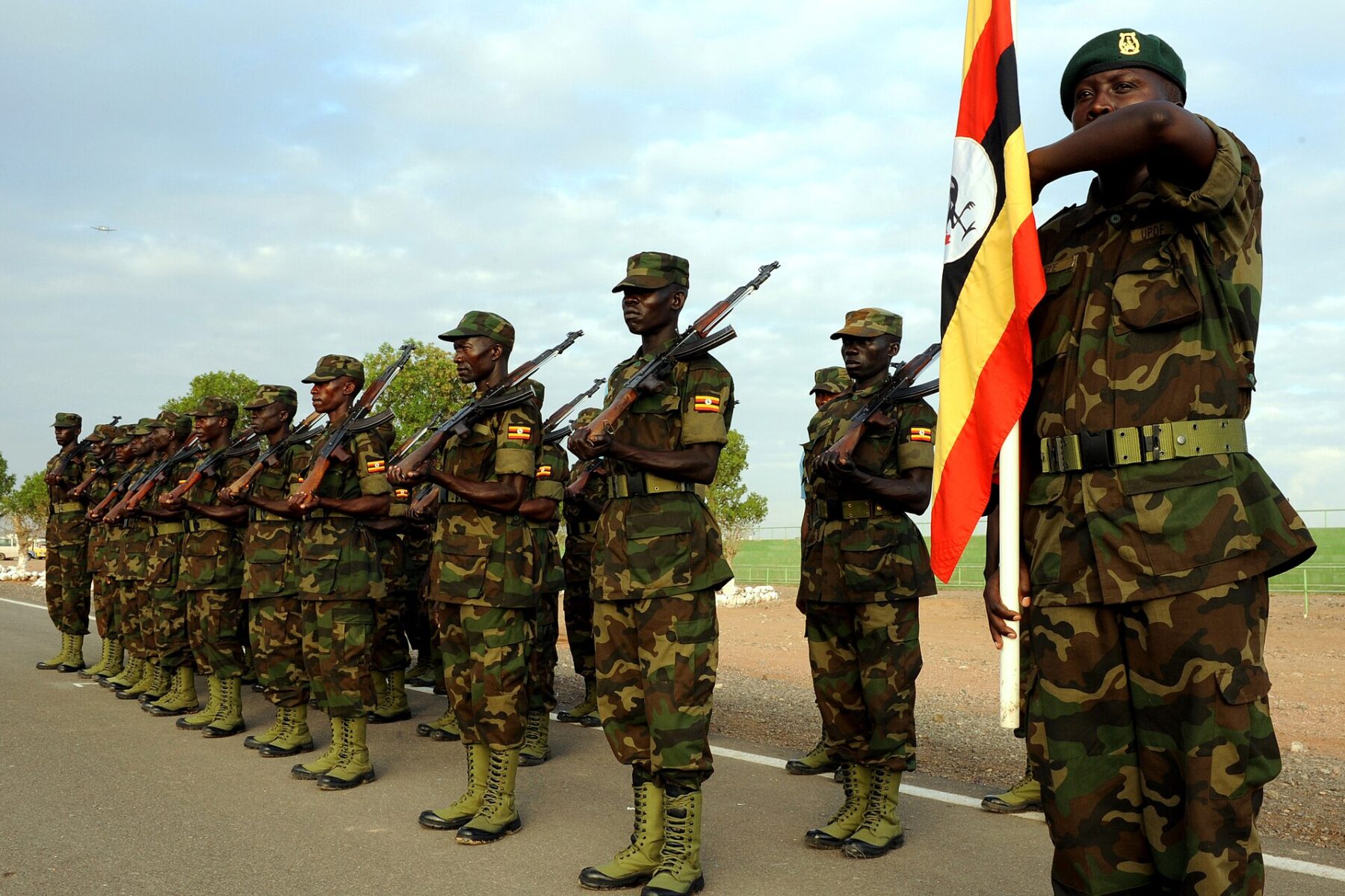 A line of Ugandan soldiers in camouflage uniforms stands to attention with rifles while one soldier holds the Ugandan flag