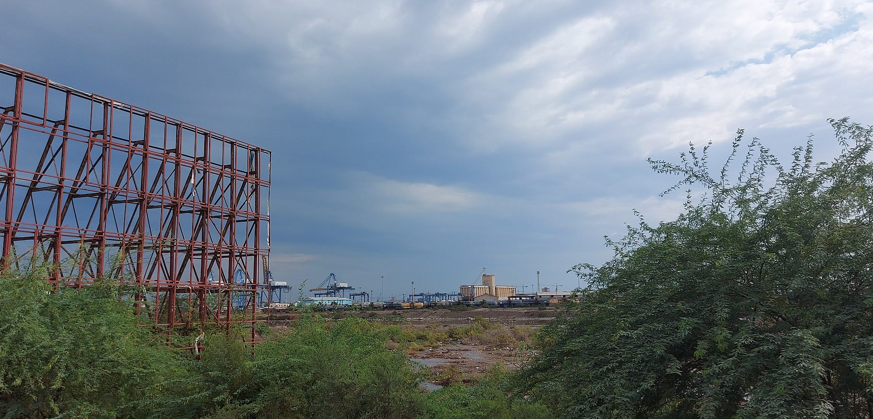 Container cranes and grain silos at the port of Port Sudan seen through trees and an empty billboard frame