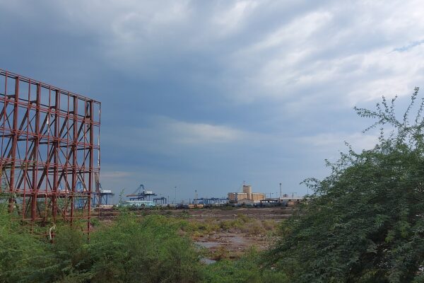 Container cranes and grain silos at the port of Port Sudan seen through trees and an empty billboard frame