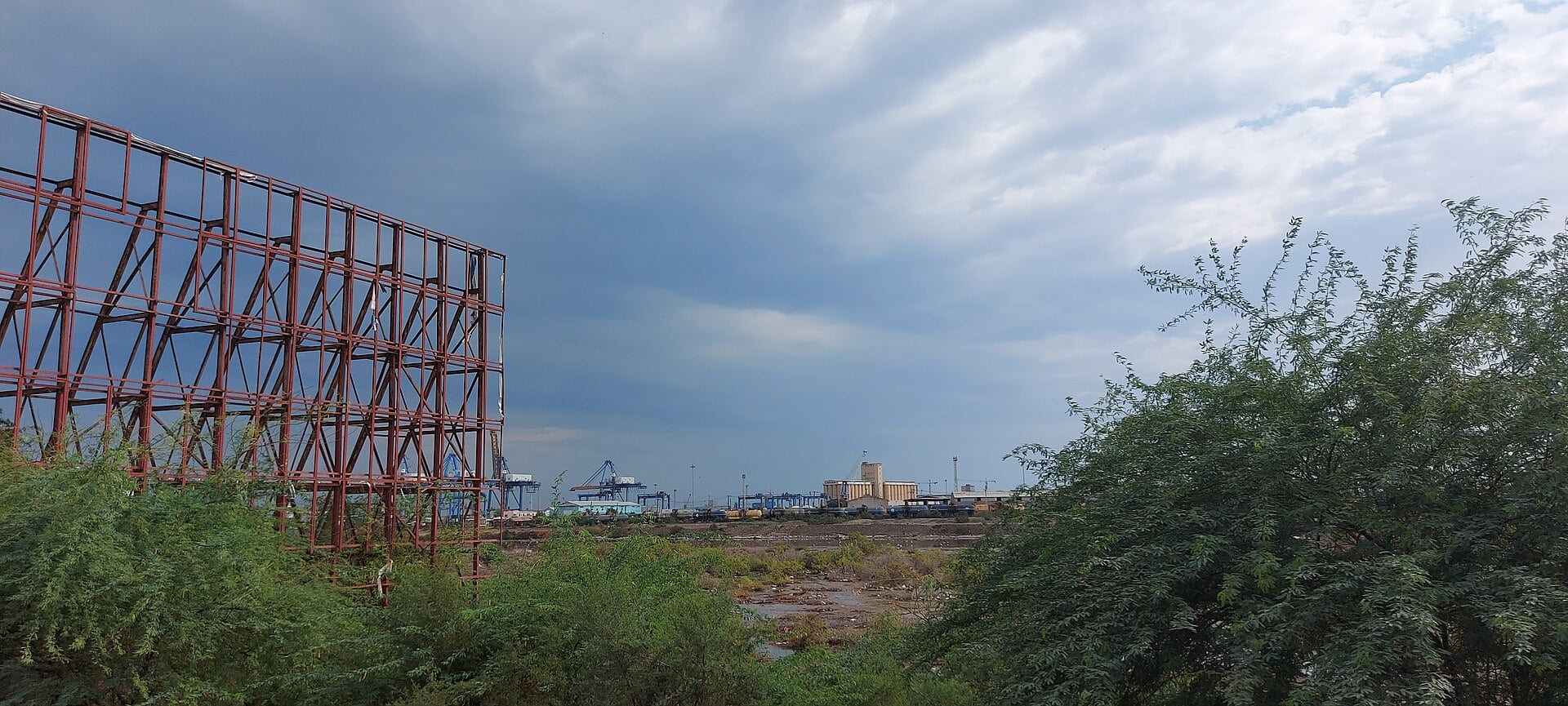 Container cranes and grain silos at the port of Port Sudan seen through trees and an empty billboard frame
