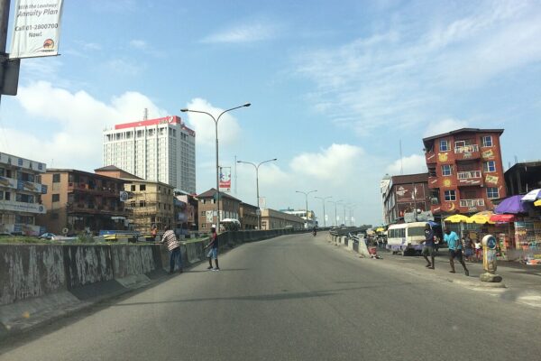 Elevated road in Lagos with the white UBA House tower rising behind low-rise buildings and street traders
