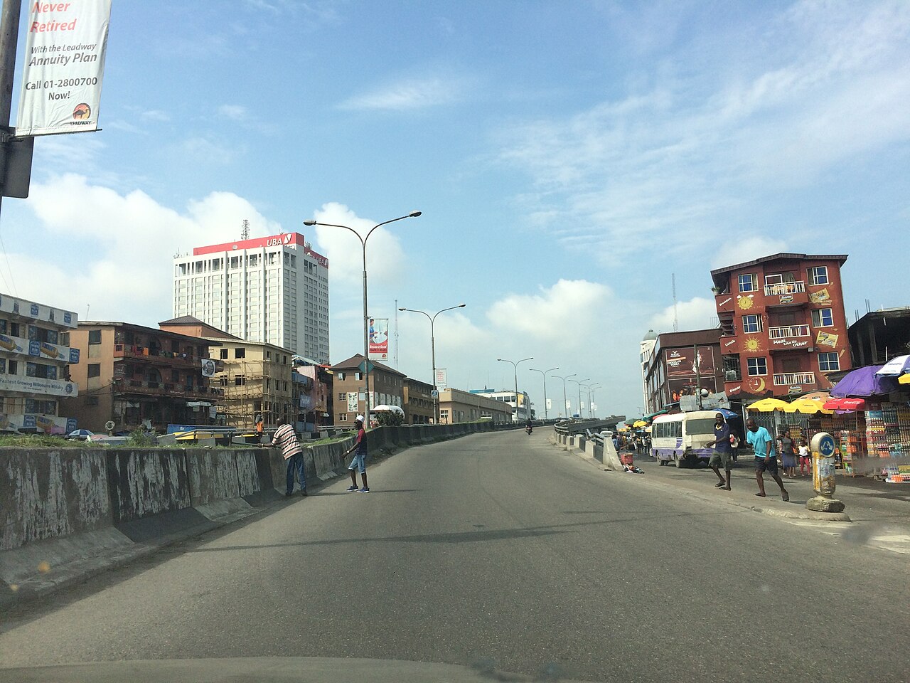 Elevated road in Lagos with the white UBA House tower rising behind low-rise buildings and street traders