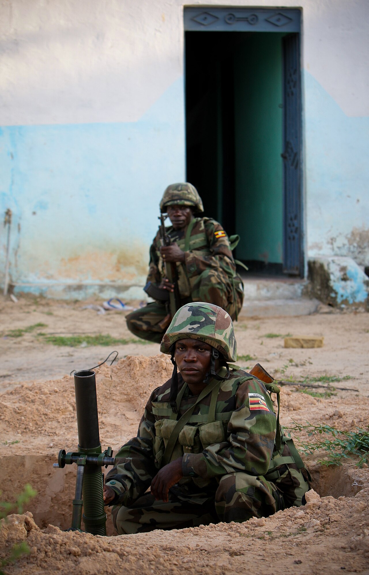Two Ugandan soldiers in camouflage and helmets at a mortar position dug into sandy ground beside a blue and white wall