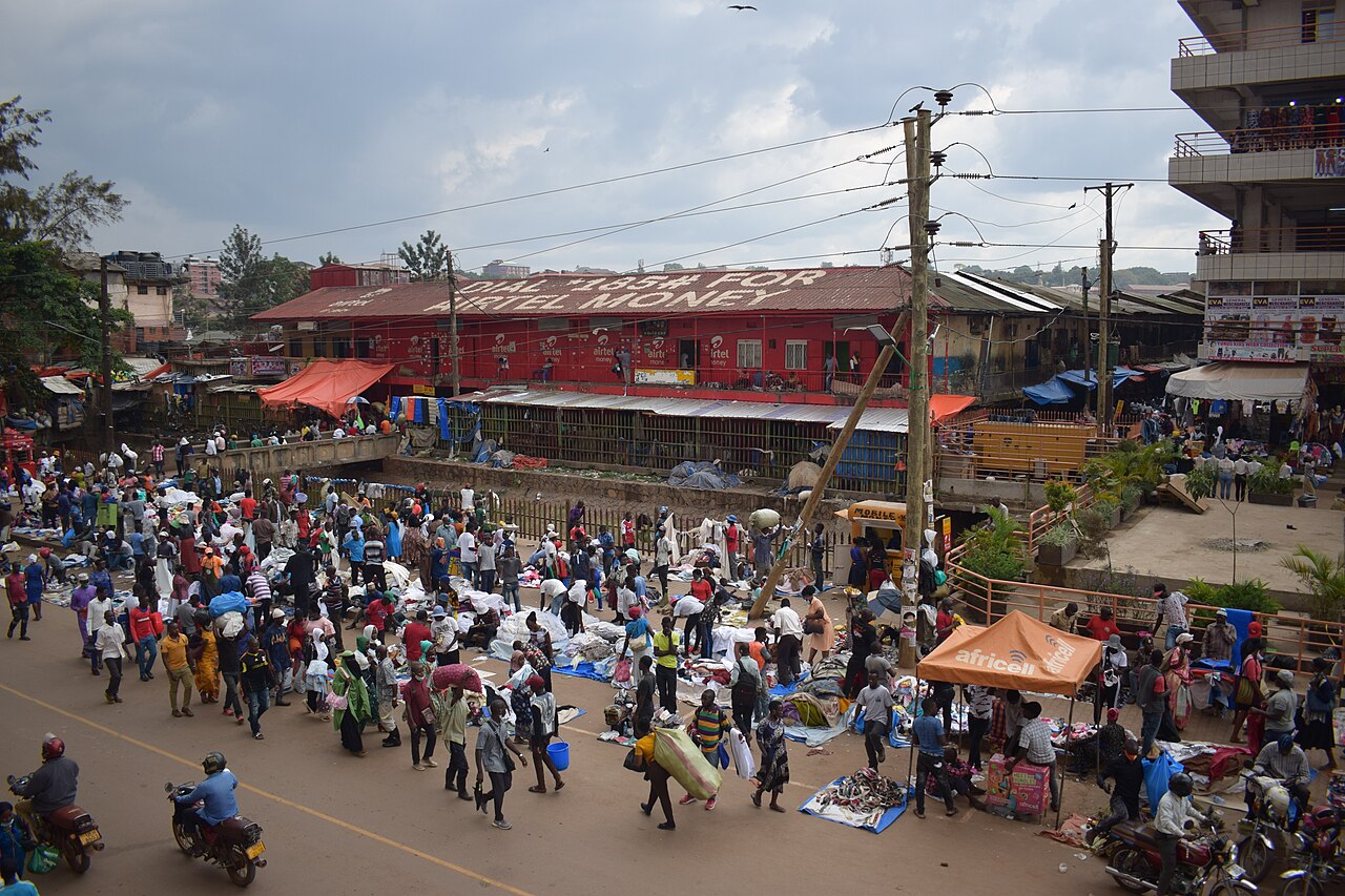 Traders and shoppers crowd the street outside Owino Market in central Kampala, with goods laid out on the ground and motorcycles passing
