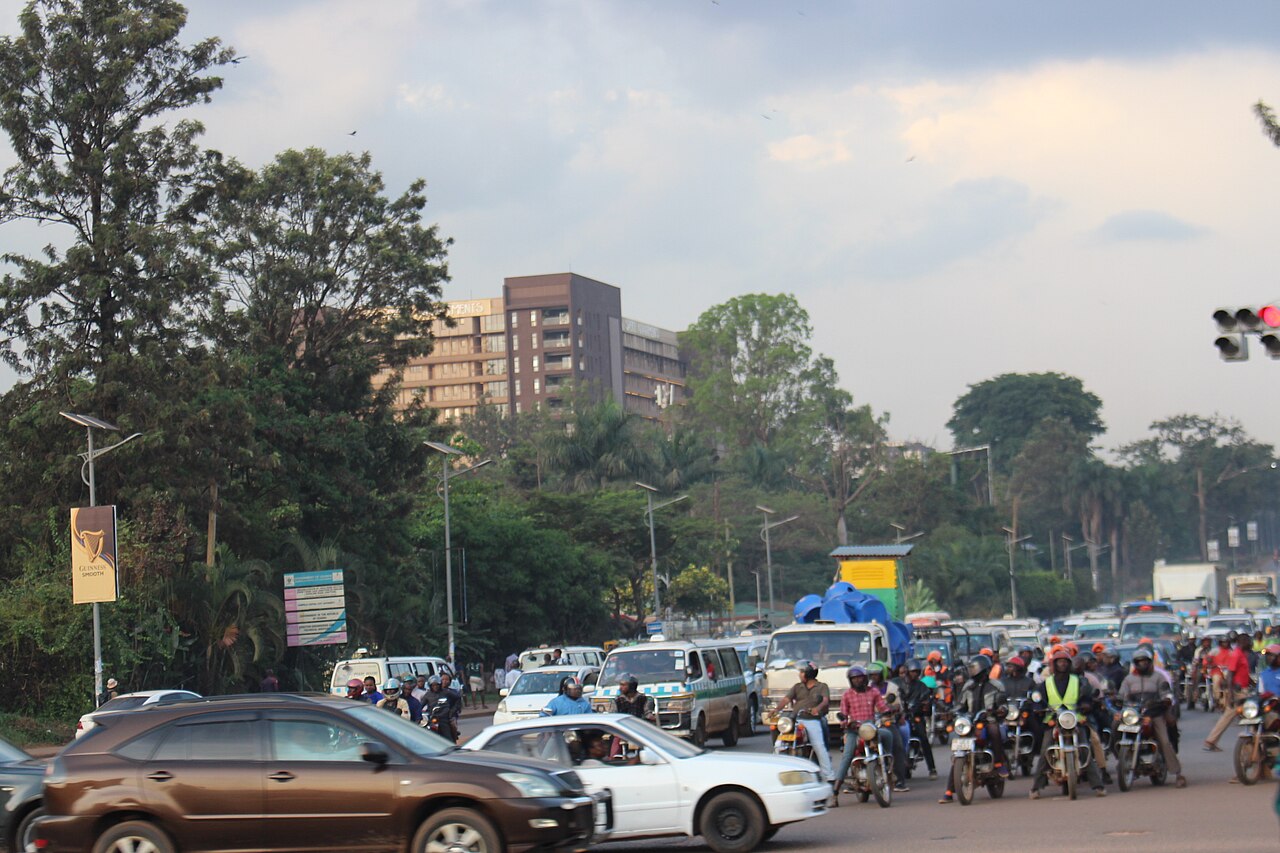 Cars, minibus taxis and motorcycle taxis waiting at a junction on Jinja Road in Kampala, with an office tower behind trees