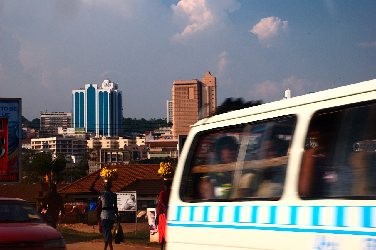 A minibus taxi and women carrying bananas on a Kampala street with office towers behind