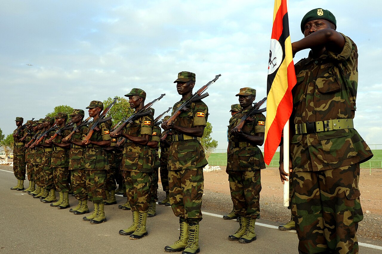 Rows of Ugandan soldiers in camouflage stand in formation with rifles beside the Ugandan flag