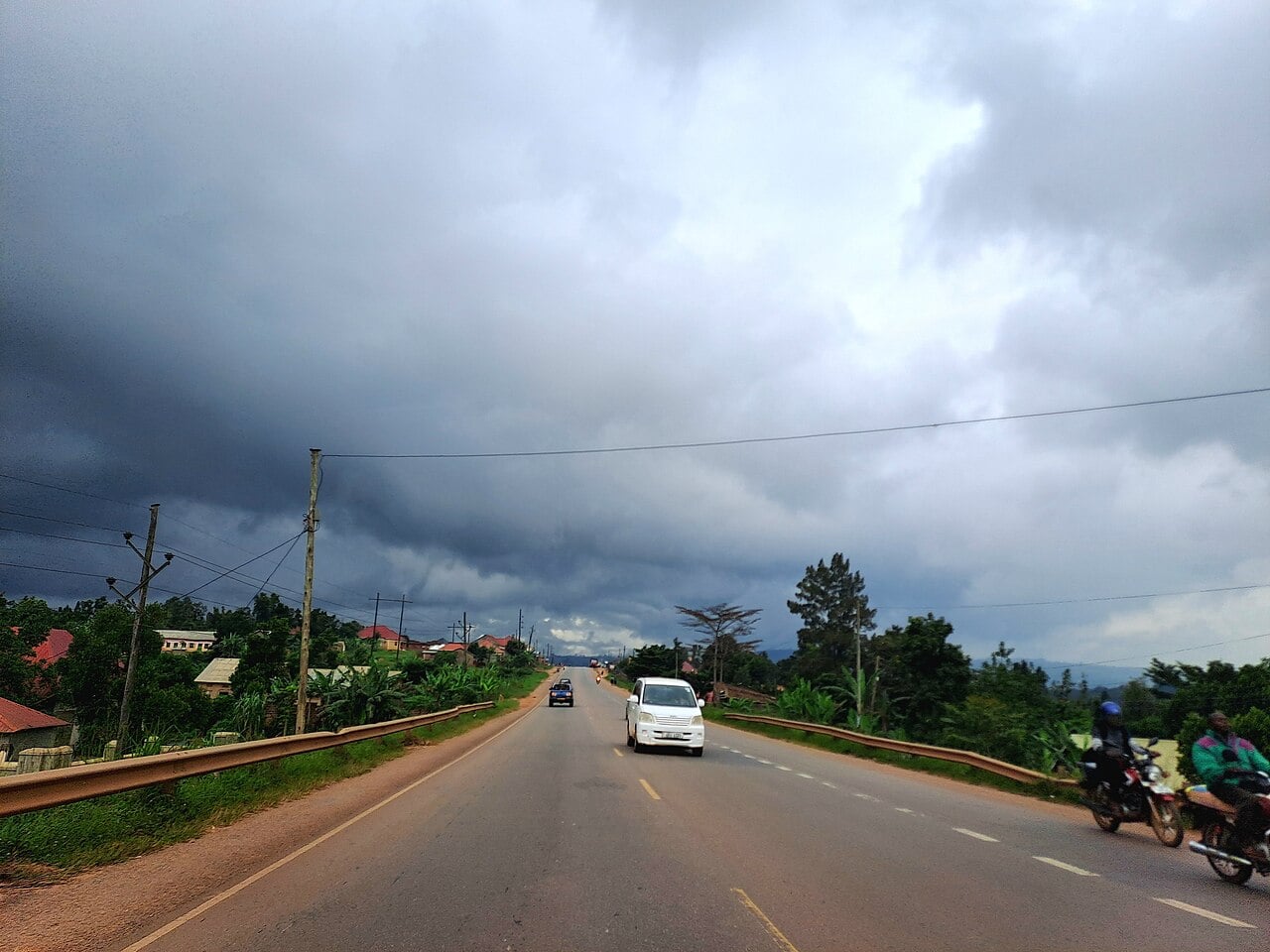 A road in Mpigi district, Uganda, under dark storm clouds, with a car and motorcycles