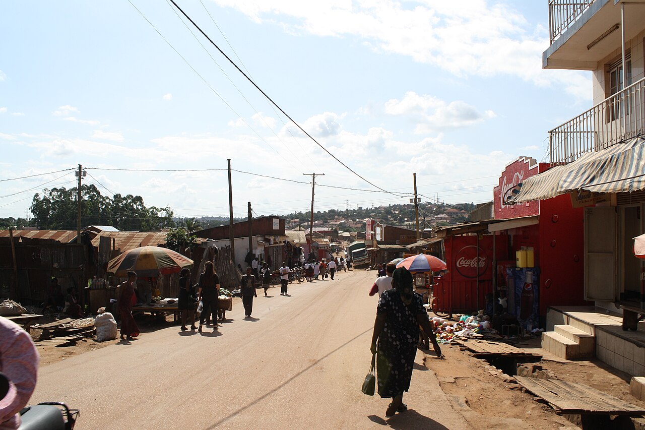 A dirt-edged main road in Kamwokya, Kampala, lined with shops and market umbrellas, with people walking under power lines