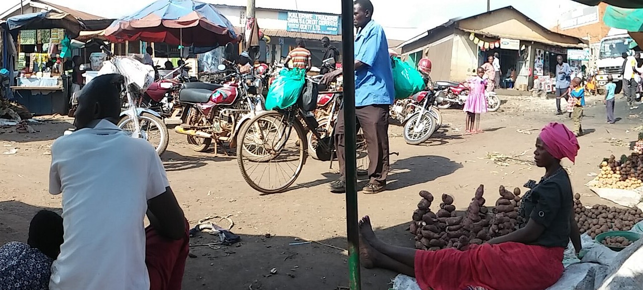 Traders, a woman selling sweet potatoes and parked motorcycle taxis at Kalerwe Market in Kampala