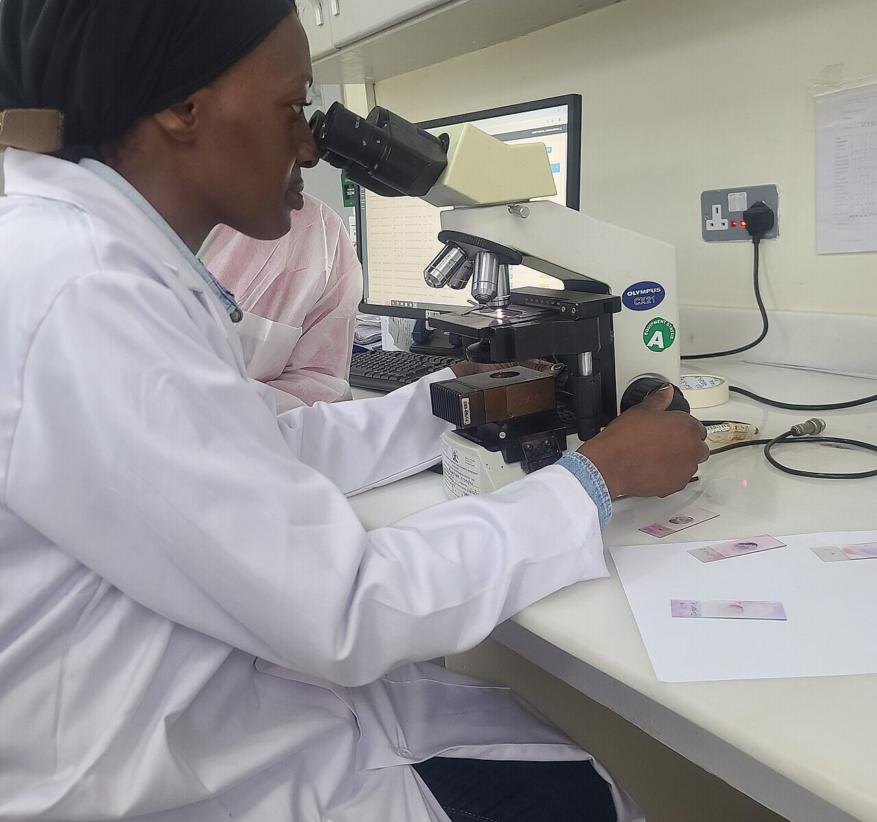 A health worker in a white coat looks through a microscope at blood slides in a laboratory at a Ugandan health facility