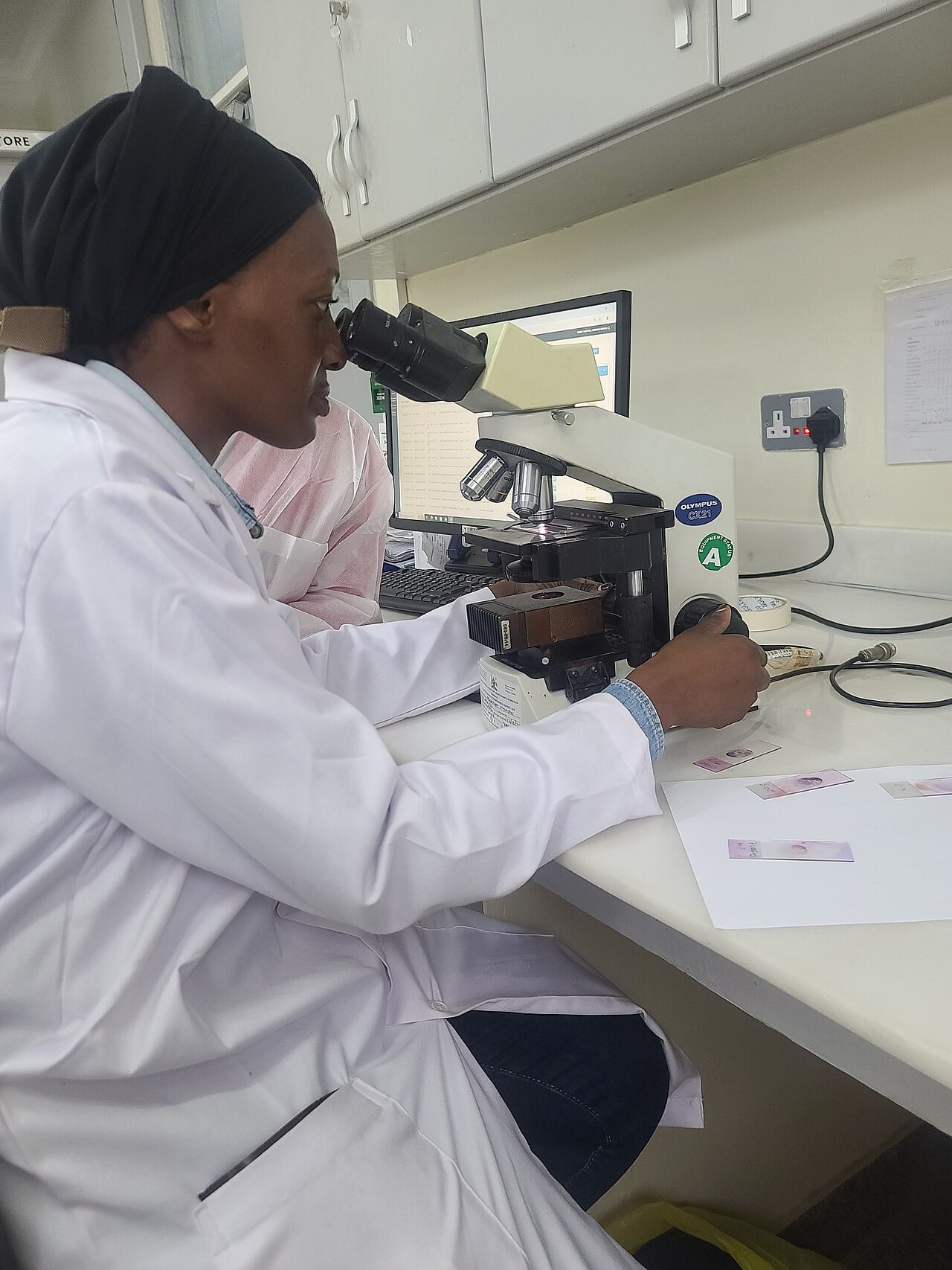A health worker in a white coat looks through a microscope at slides in a laboratory at a Ugandan health facility