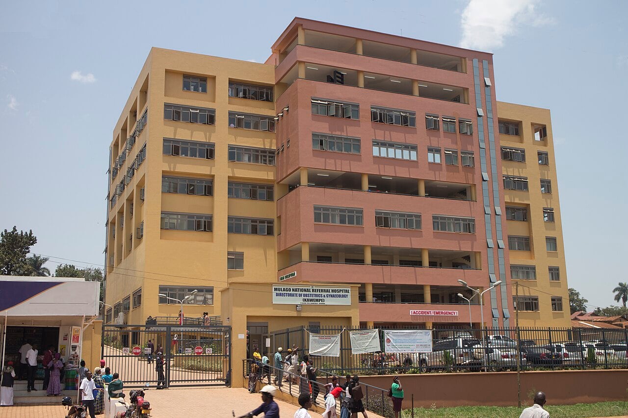 A multi-storey yellow and pink public hospital building in Kampala, Uganda, with people walking at its entrance