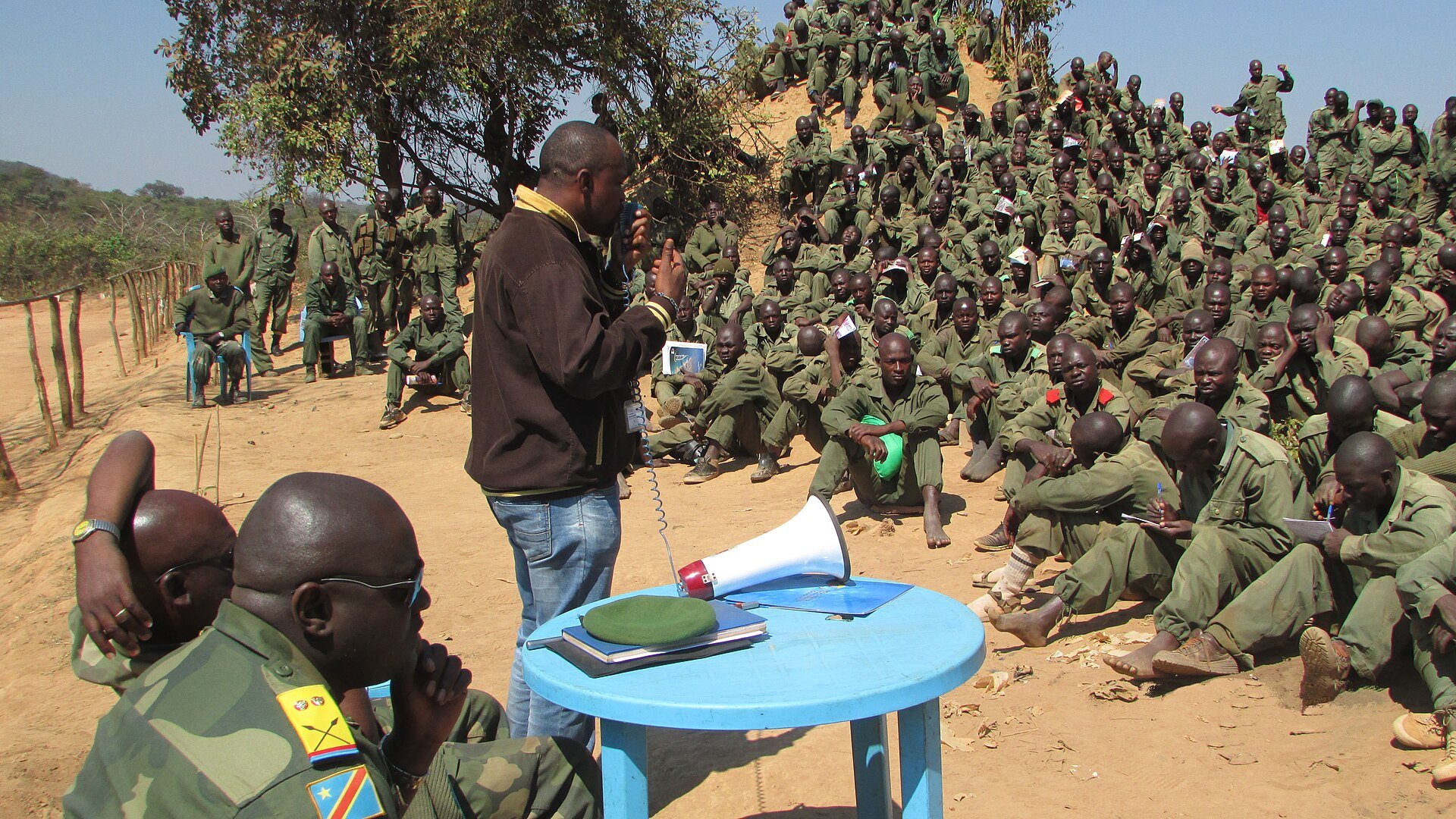 A man in civilian clothes with a megaphone speaks to hundreds of seated Congolese army soldiers on a hillside while officers sit in the foreground