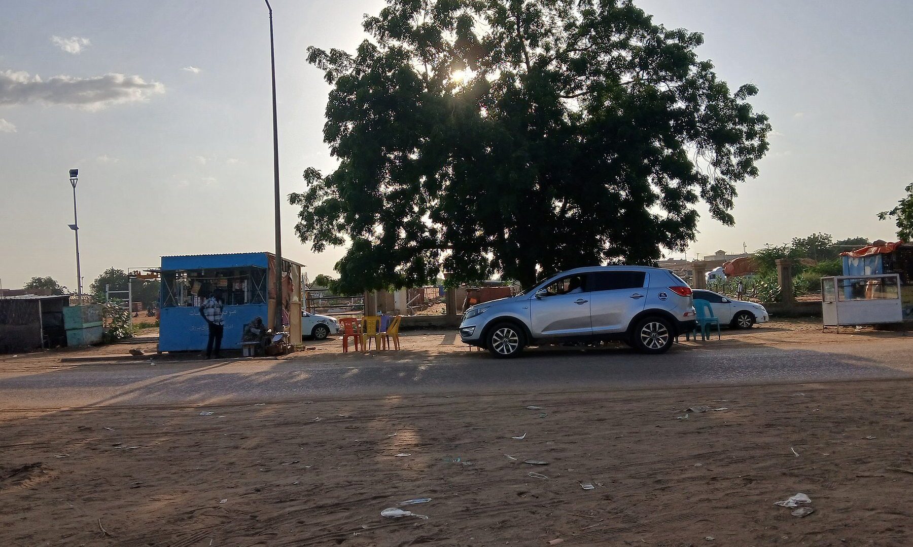 A street in El Obeid, Sudan, with roadside stalls, parked cars and a large tree at dusk