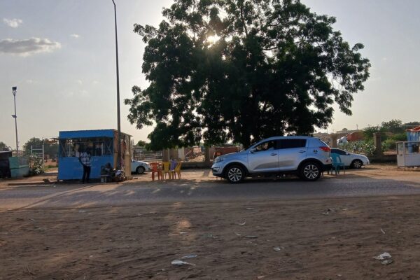 A street in El Obeid, Sudan, with roadside stalls, parked cars and a large tree at dusk