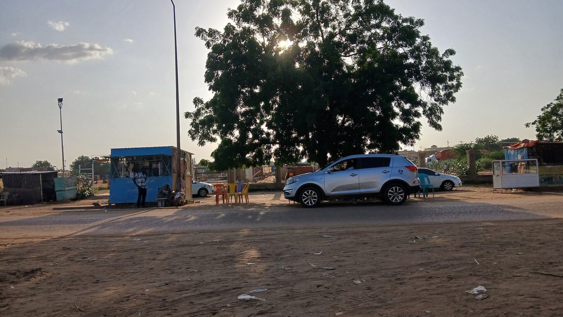 A street in El Obeid, Sudan, with roadside stalls, parked cars and a large tree at dusk