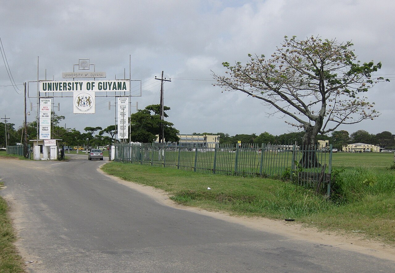 The arched gateway of the University of Guyana with its coat of arms and faculty signs, a paved road, a car and a green fenced field under a cloudy sky