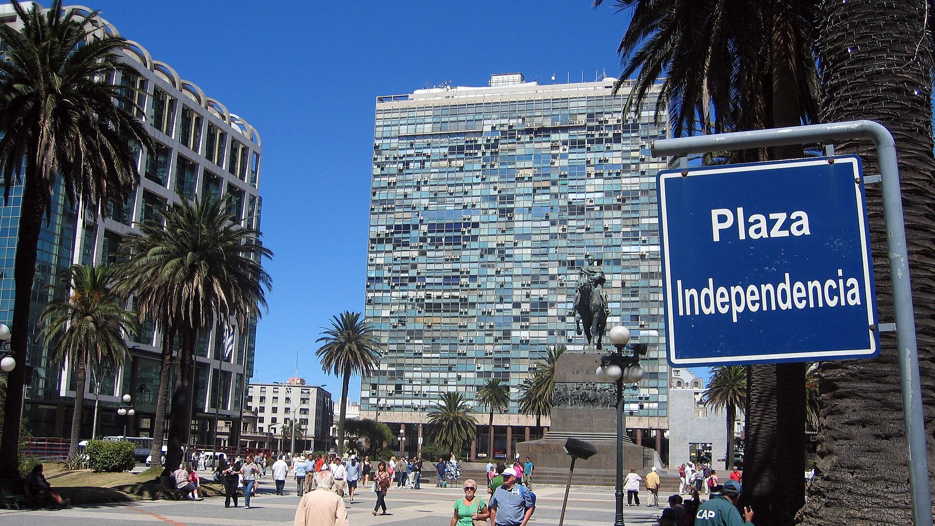 Pedestrians cross Plaza Independencia in Montevideo under palm trees, with the Artigas statue and a tower block behind