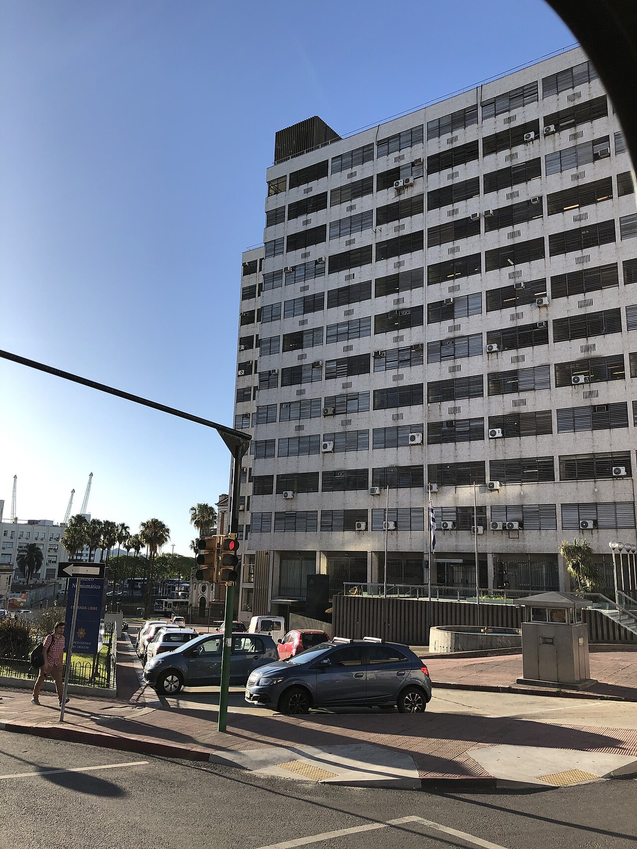 A white multi-storey office block with rows of window blinds and a Uruguayan flag at the entrance, with cars parked in front.