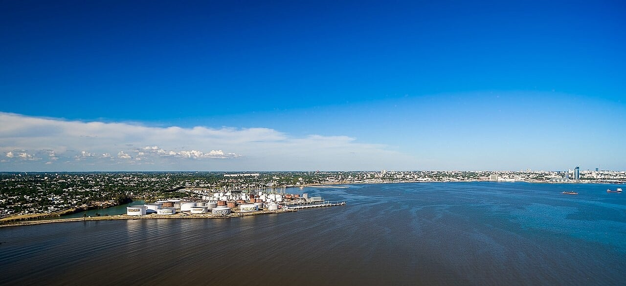 Aerial view of the La Teja oil refinery tank farm on Montevideo Bay, with the Montevideo skyline across the water