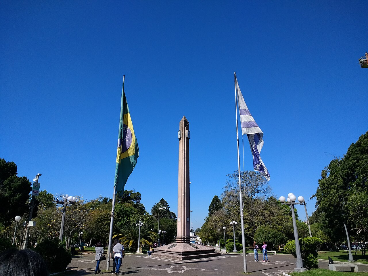 Brazilian and Uruguayan flags fly either side of a tall obelisk in the Plaza Internacional, with pedestrians walking across the square